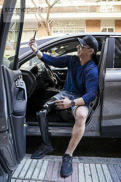 Lächelnder Mann mit Beinprothese  der im Auto ein Selfie mit dem Smartphone macht