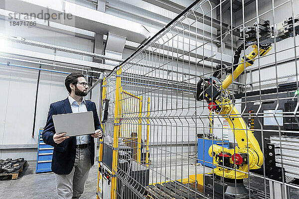 Junger Geschäftsmann mit Laptop analysiert Roboterarm in Fabrik