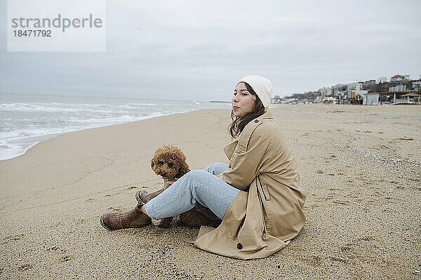 Junge Frau mit Maltipoo-Hund sitzt auf Sand am Strand