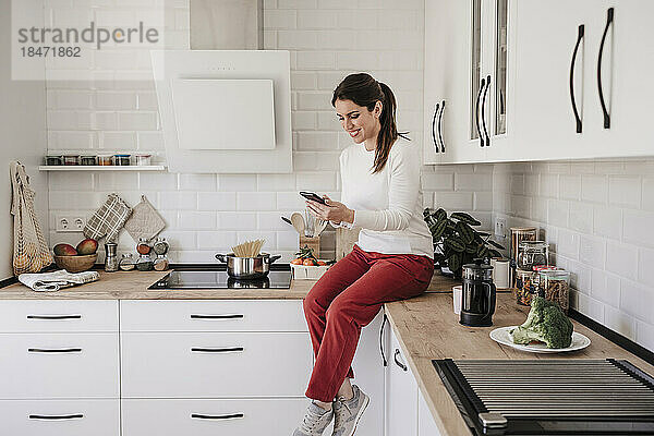 Lächelnde Frau  die ihr Smartphone benutzt und auf der Theke in der Küche sitzt