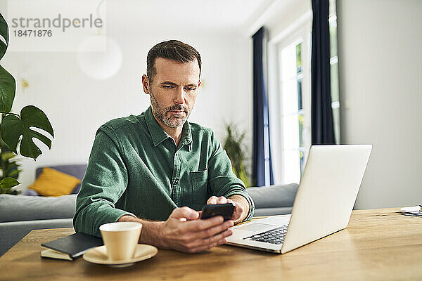 Mann benutzt Telefon  während er von zu Hause aus arbeitet und am Tisch mit Laptop sitzt