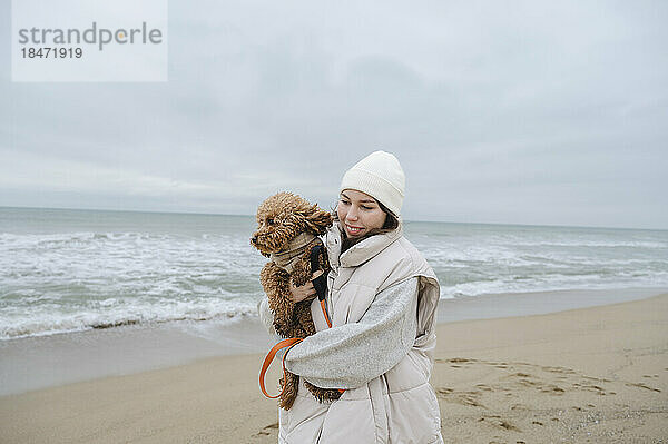 Smiling woman wearing warm clothing holding pet dog at beach
