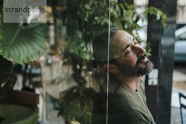 Bearded mature man with eyes closed leaning on glass wall in cafe