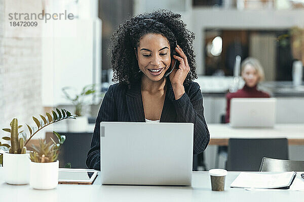 Glückliche Geschäftsfrau  die im Büro über Headset spricht und Laptop benutzt