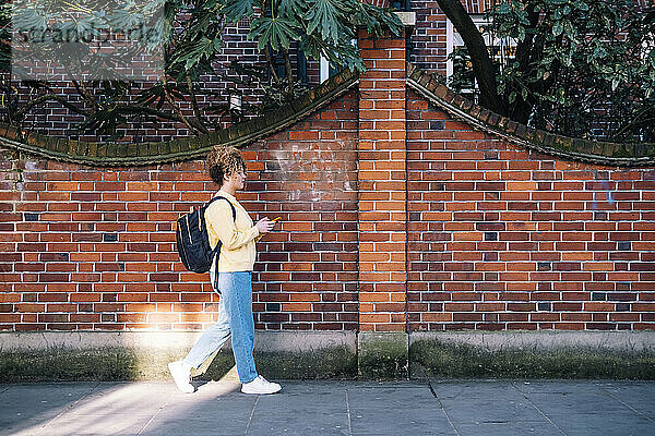 Junge Frau benutzt Smartphone und geht auf Fußweg an Ziegelmauer entlang
