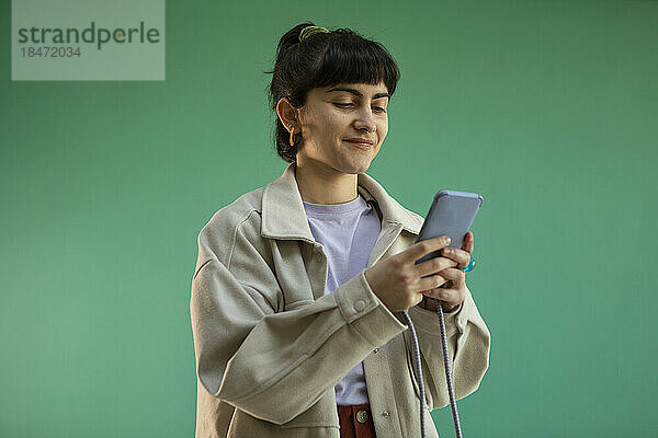 Lächelnde junge Frau  die ihr Mobiltelefon vor grünem Hintergrund benutzt