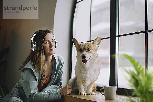 Nachdenkliche Frau mit Kopfhörern sitzt zu Hause mit Hund am Fenster