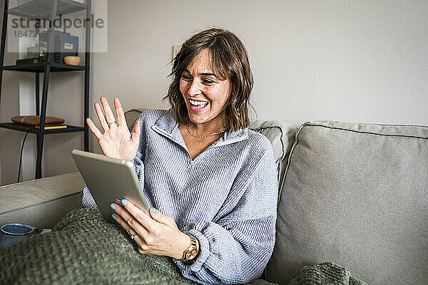 Glückliche Frau winkt per Videoanruf durch den Tablet-PC auf dem heimischen Sofa