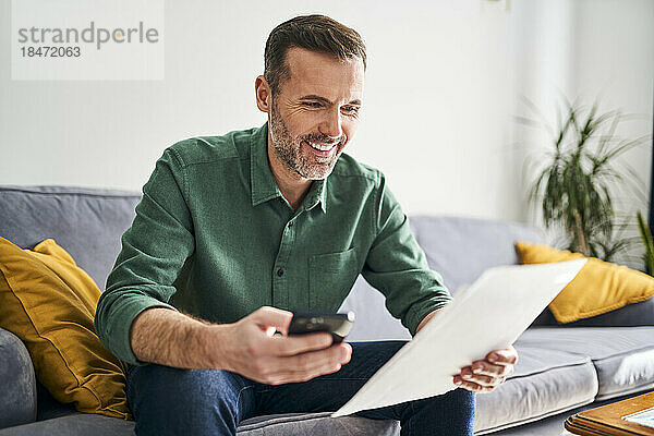 Lächelnder Mann sitzt auf der Couch und bezahlt Rechnungen mit seinem Smartphone