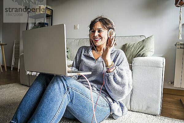 Glückliche Frau winkt durch ihren Laptop und sitzt zu Hause auf dem Boden