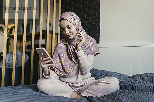 Lächelnde junge Frau mit Hijab  die im Schlafzimmer Textnachrichten per Smartphone sendet