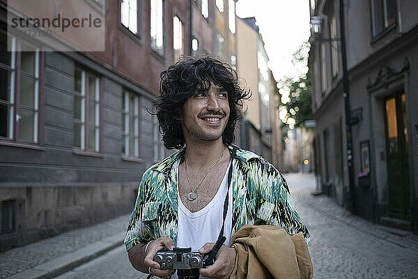 Lächelnder Mann steht mit Kamera inmitten von Gebäuden