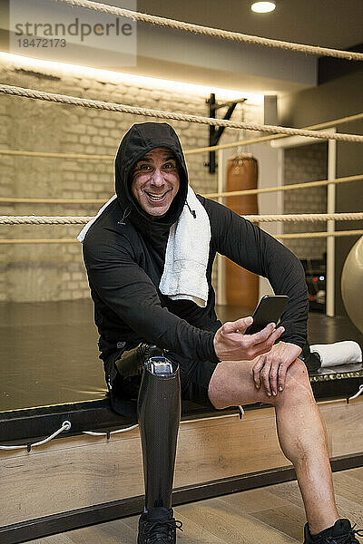 Glücklicher Mann mit Behinderung sitzt auf Boxring und hält Smartphone im Fitnessstudio