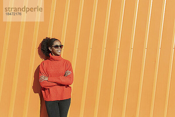 Glückliche Frau mit Sonnenbrille vor orangefarbener Wand an einem sonnigen Tag