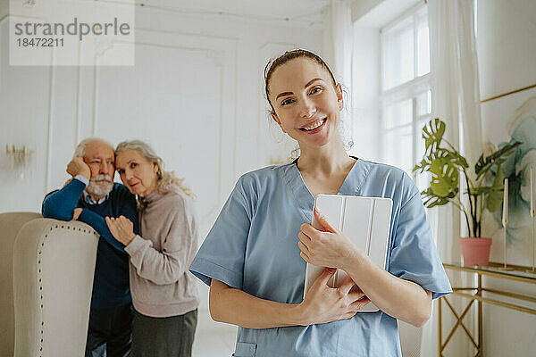 Lächelnde Krankenschwester hält Tablet-PC mit älterem Mann und älterer Frau im Hintergrund zu Hause