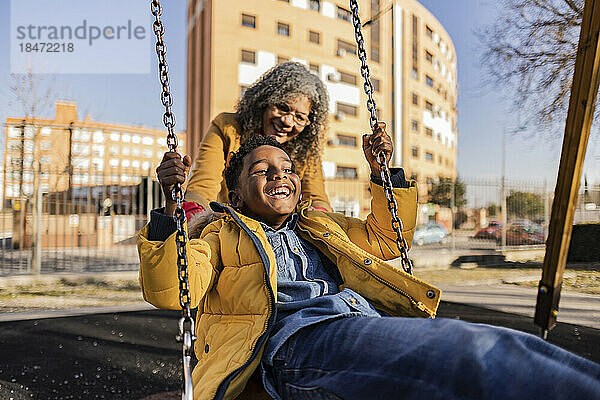 Verspielte Seniorin schiebt ihren glücklichen Enkel auf der Schaukel auf dem Spielplatz