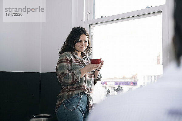 Lächelnde Geschäftsfrau mit Kaffeetasse steht am Fenster im Büro