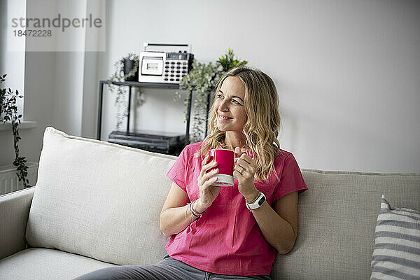 Lächelnde reife Frau mit Kaffeetasse sitzt zu Hause auf dem Sofa
