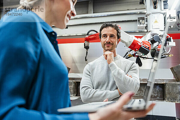 Lächelnder Ingenieur mit der Hand am Kinn und Blick auf Kollegen in der Roboterfabrik
