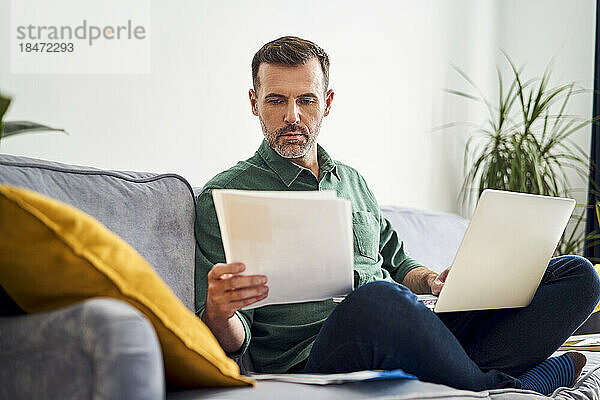 Besorgter Mann  der von zu Hause aus arbeitet und Dokumente betrachtet  während er mit einem Laptop auf dem Sofa sitzt