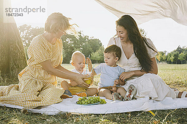 Glückliche Frauen sitzen mit Söhnen beim Picknick und halten ein Glas Saft in der Hand