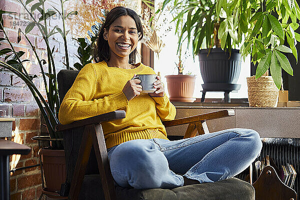 Glückliche Frau hält eine Kaffeetasse in der Hand und sitzt mit gekreuzten Beinen im Stuhl zu Hause
