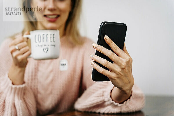 Lächelnde Frau benutzt Smartphone und trinkt zu Hause Kaffee