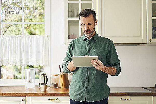 Reifer Mann mit digitalem Tablet  der in der Küche steht
