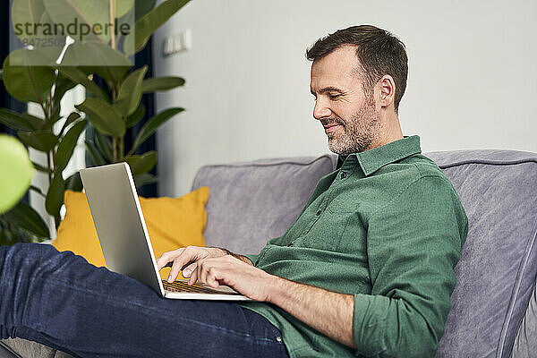 Entspannter Mann  der von zu Hause aus mit einem Laptop auf dem Sofa arbeitet