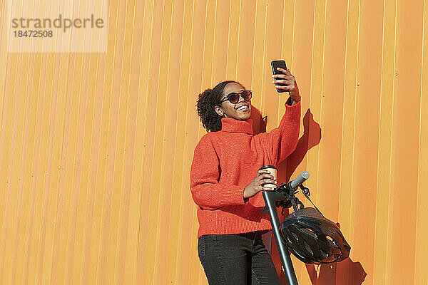 Frau macht Selfie per Smartphone mit elektrischem Tretroller  der an orangefarbener Wand steht