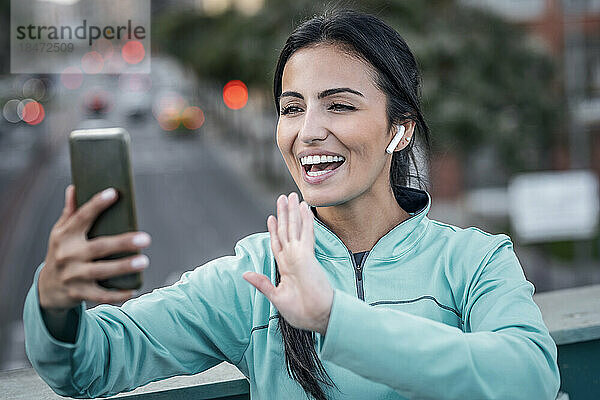 Glückliche junge Frau winkt bei einem Videoanruf über ihr Mobiltelefon