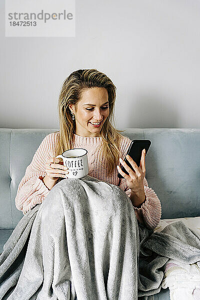 Lächelnde junge Frau  die zu Hause ihr Smartphone mit einer Kaffeetasse benutzt