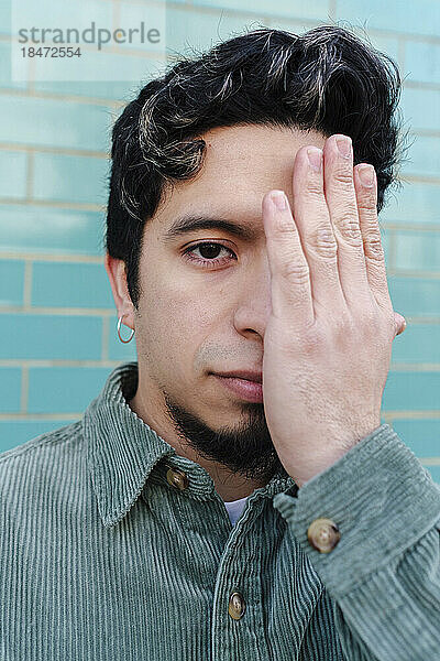Man covering eye with hand in front of turquoise brick wall