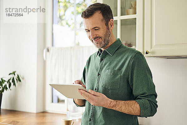 Reifer Mann mit digitalem Tablet  der in der Küche steht