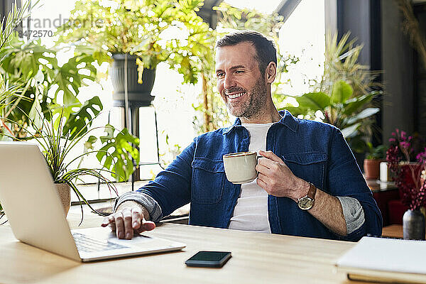Glücklicher Geschäftsmann benutzt Laptop und trinkt Kaffee im Loft-Büro