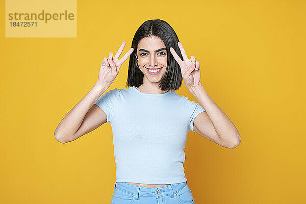 Smiling young woman gesturing peace sign against yellow background