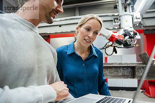 Reifer Ingenieur blickt auf Laptop  der von einem Kollegen in der Roboterfabrik gehalten wird