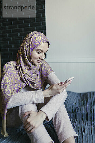 Lächelnde junge Frau mit Hijab und Smartphone  die zu Hause im Bett sitzt