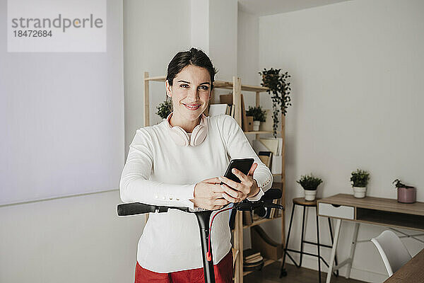 Lächelnde Geschäftsfrau steht mit Elektroroller und hält Smartphone im Büro