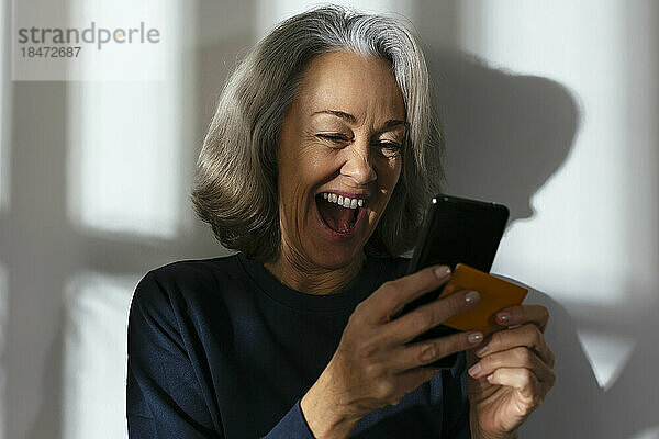 Fröhliche reife Frau mit Kreditkarte und Smartphone vor der Wand