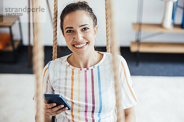 Glückliche junge Geschäftsfrau mit Mobiltelefon sitzt im Büro