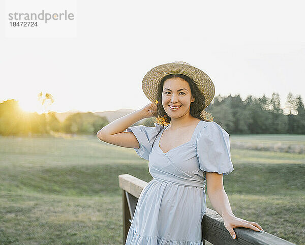 Lächelnde Frau mit Hut steht bei Sonnenuntergang am Geländer