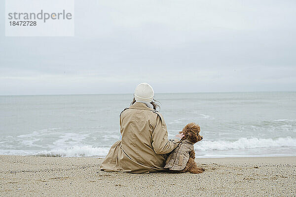 Frau sitzt mit Hund am Ufer und schaut aufs Meer