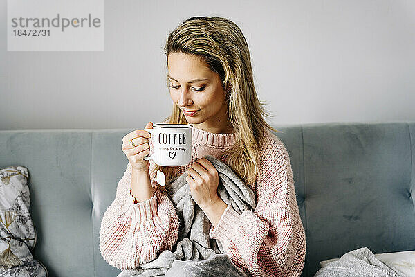 Lächelnde junge Frau trinkt Kaffee und sitzt zu Hause auf dem Sofa