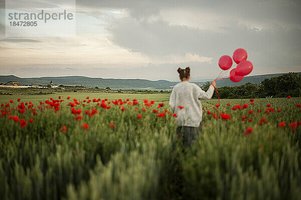Mädchen läuft bei Sonnenuntergang mit roten Luftballons im Mohnfeld