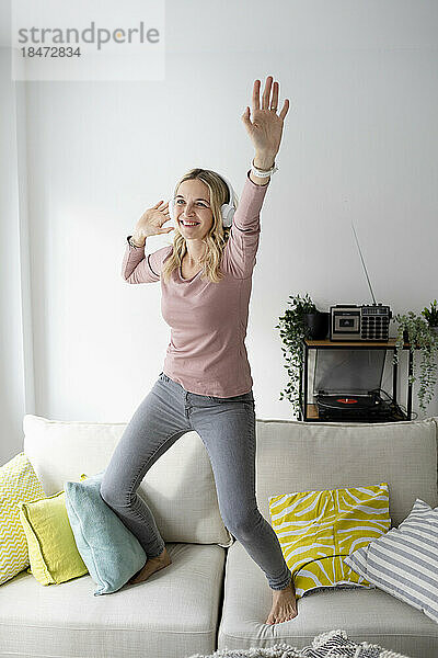 Happy woman wearing wireless headphones listening to music and dancing on sofa at home