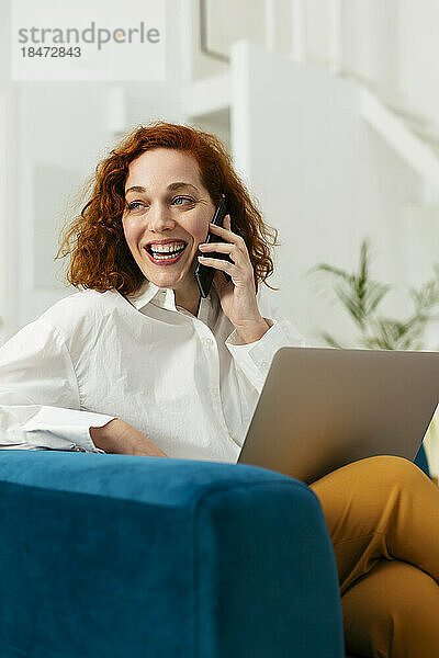 Glückliche Geschäftsfrau mit Rotschopf  die im Büro auf dem Smartphone spricht