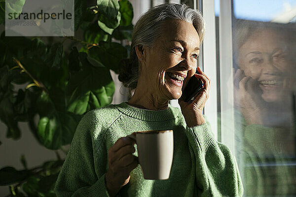 Glückliche reife Frau  die zu Hause am Fenster mit dem Handy telefoniert