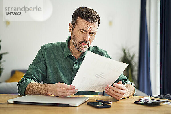 Verärgerter Mann schaut sich Dokument mit Finanzzahlen an  während er zu Hause am Tisch sitzt