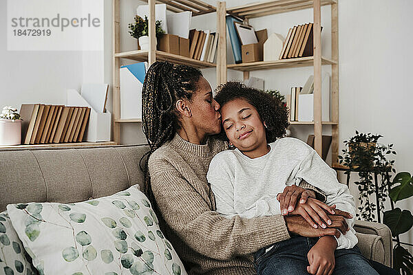 Mutter küsst Tochter  die zu Hause auf dem Sofa sitzt